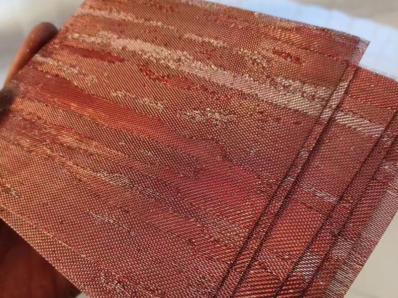 Fine woven metal fabric in copper tones, held up to the light, showing the crimped weave